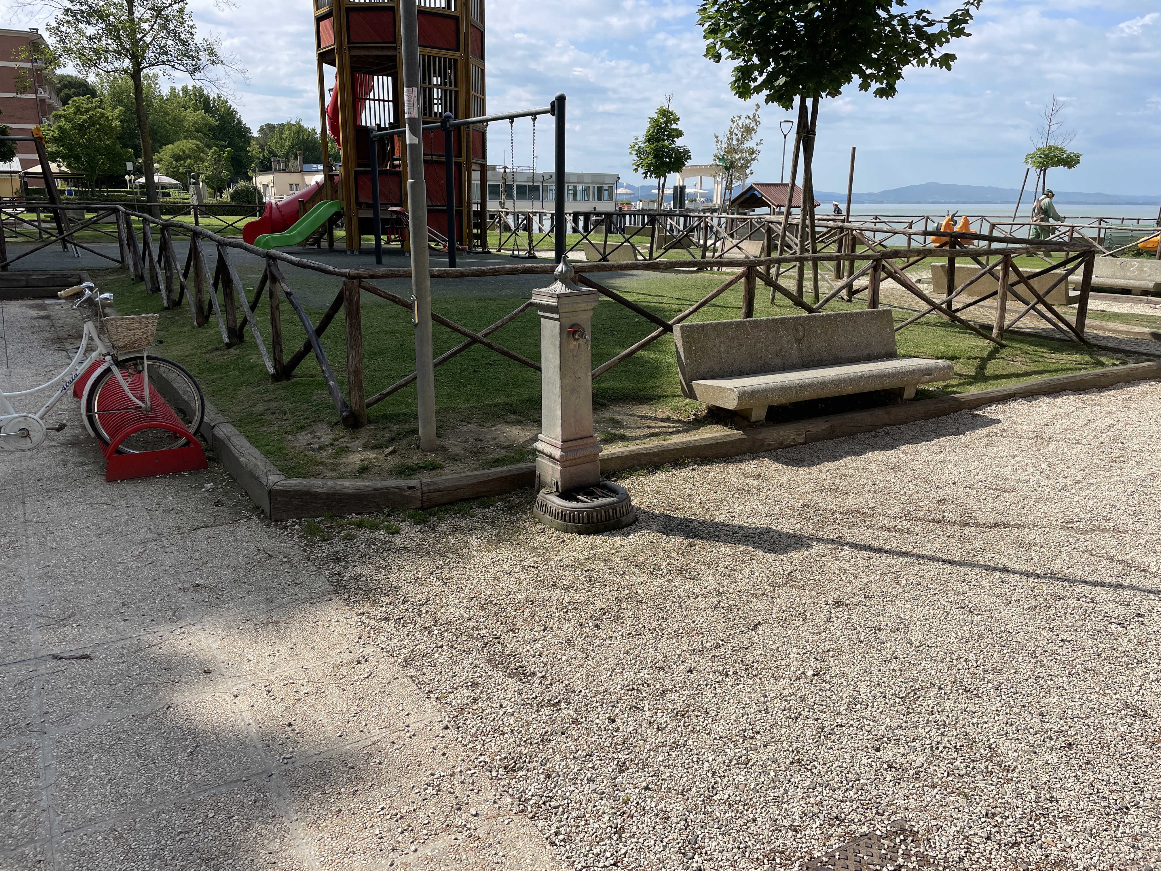 Passignano playground area. At left a white bike. Bench and drinking fountain at the entrance to the fenced playground.